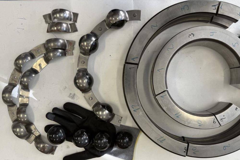 Disassembled and cut sections of a large wind turbine bearing laid out on a white surface. On the right are two concentric bearing rings cut into numbered segments. On the left are rows of metal ball retainers holding shiny steel balls, alongside four loose bearing balls resting on a black work glove.